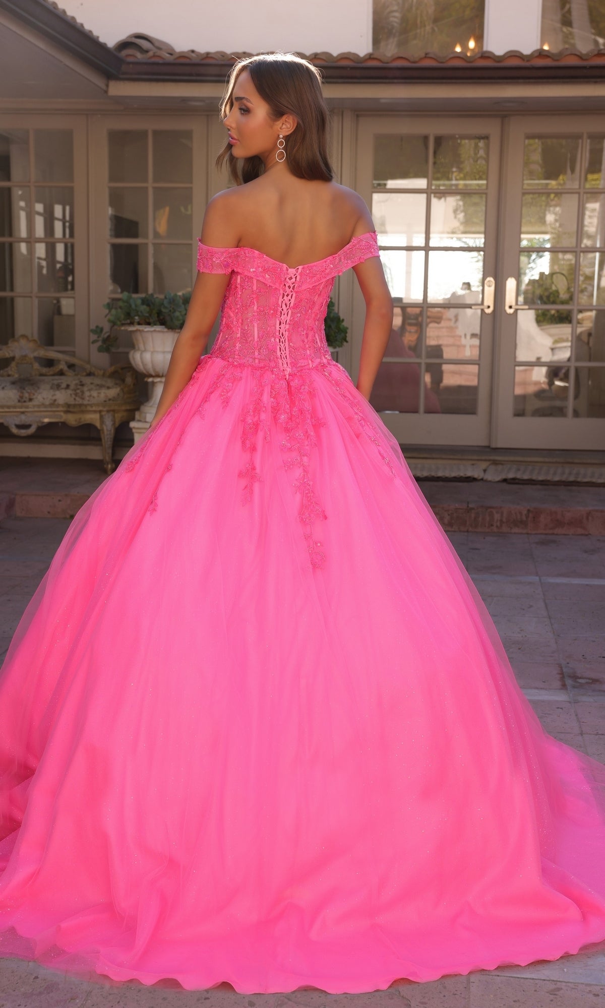 A woman stands outdoors facing away, wearing the Bright Off-the-Shoulder Long Prom Ball Gown H1349 in vivid pink with floral details and a full skirt. French doors and a patio serve as the elegant backdrop for this stunning formal dress.